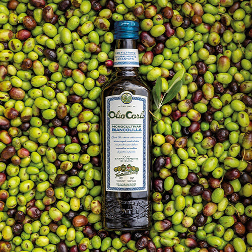 Bottiglia di Olio Carli Extra Vergine di Oliva Monocultivar Biancolilla adagiata su un letto di olive.