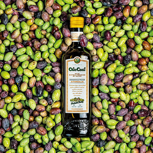 Bottiglia di Olio Carli Extra Vergine di Oliva Monocultivar Athinolia adagiata su un letto di olive.